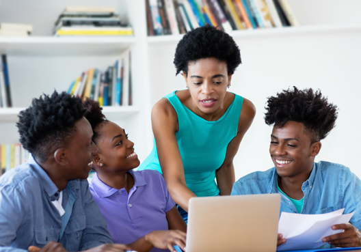 Classroom scene with a teacher and three learners Classroom scene with a teacher and three learners who are seated around a table looking at a laptop screen, bookshelves in the background